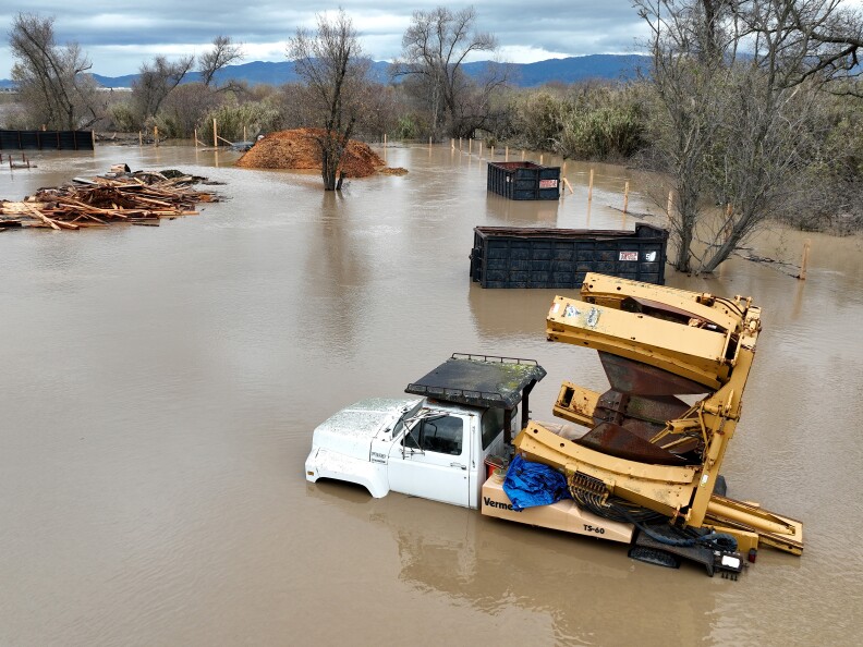 Brown water is past the tires on a truck carrying equipment. Beyond the large stretch of flooded land hills and trees are visible.