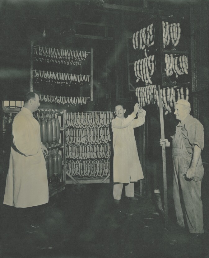 A black and white photo of Vienna Beef workers inspecting sausages at the Chicago factory. Sausages are hanging in rows everywhere around them