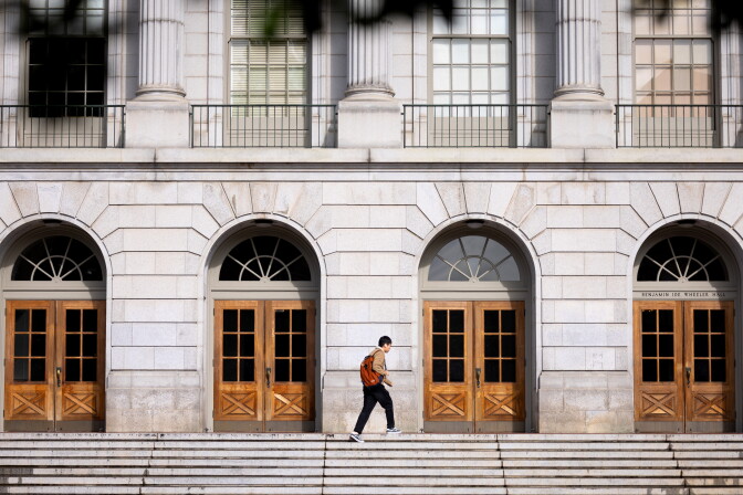 A person with short black hair and light brown skin, wearing a tan jacket, black pants, and black-and-white sneakers, walks in profile along a stone step with a rust-orange backpack over their shoulder. Behind them, the gray granite facade of Benjamin Ide Wheeler Hall has four arched entryways, each with a fan-shaped transom window and a pair of wooden double doors with glass panes.