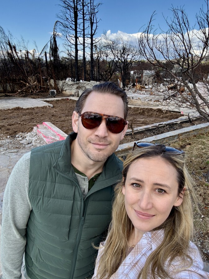 A younger middle aged couple with light skin tone takes a selfie. The woman on the right has long blond hair, and her husband, wears sunglasses and a green vest. Behind them is the rubble of their burned home. 