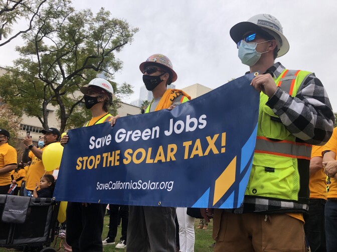 Three men wearing masks and hard hats hold a blue sign with white and orange lettering that says "Save Green Jobs, Stop the Solar Tax!"