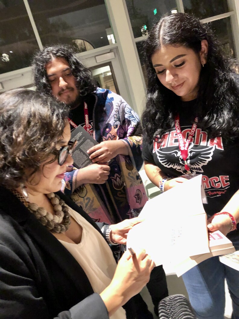 Alt text: College professor, activist and artist signs copies of her book while female college student holds it open. Student wears a t-shirt with word MEChA, the name of a Chicanx activist group.
