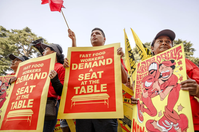 Three protesters stand side by side. Two are holding large posters that read Fast Food Workers Demand A Seat At the Table.