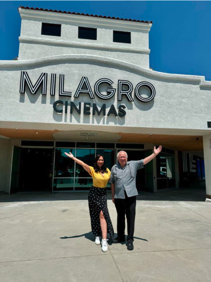 Two people raise one hand on opposite sides as they pose in front of a grey building with a sign that reads "MILAGRO CINEMAS."
