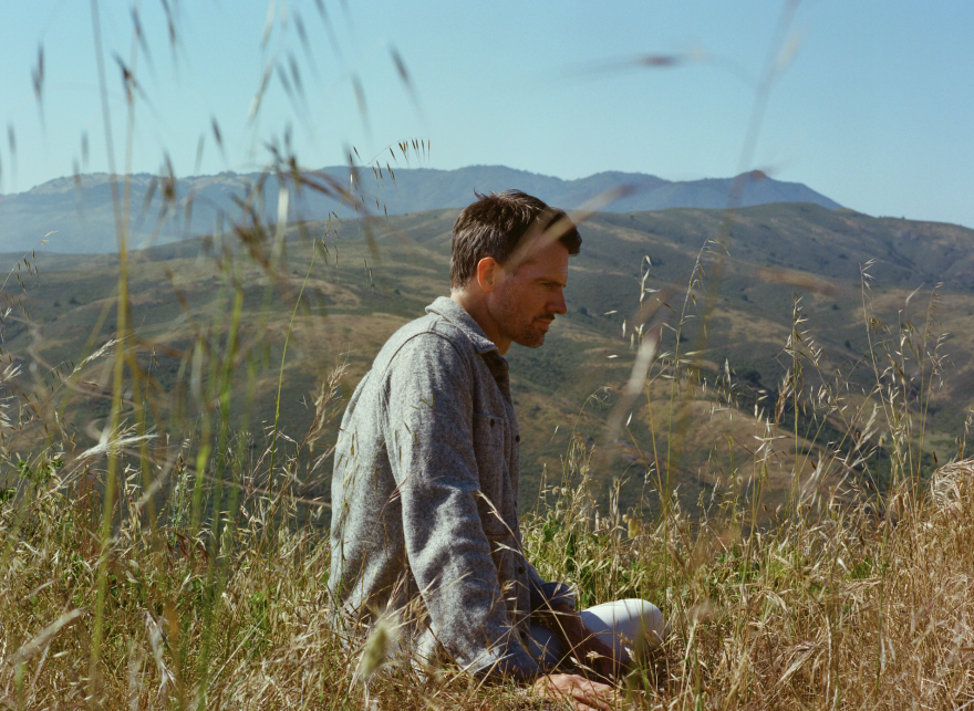 Tycho's lead singer sitting on grass with mountains in the background.