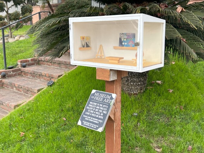 A white box with two transparent walls that showcase small works of art, including a ceramic nose and drawings. The box, located in a home's front yard, is accompanied with a sign that reads "Museum of Free Art"