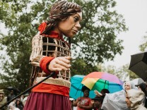Little Amal, a 12 ft. puppet towers over the audience beneath her at a park. She has a long, red, flowing skirt, and red ribbon in her braided hair. The puppet’s torso is made of an open cage inside which you can see one puppeteer inside. Her right hand is outstretched, manipulated by a long black rod. 
