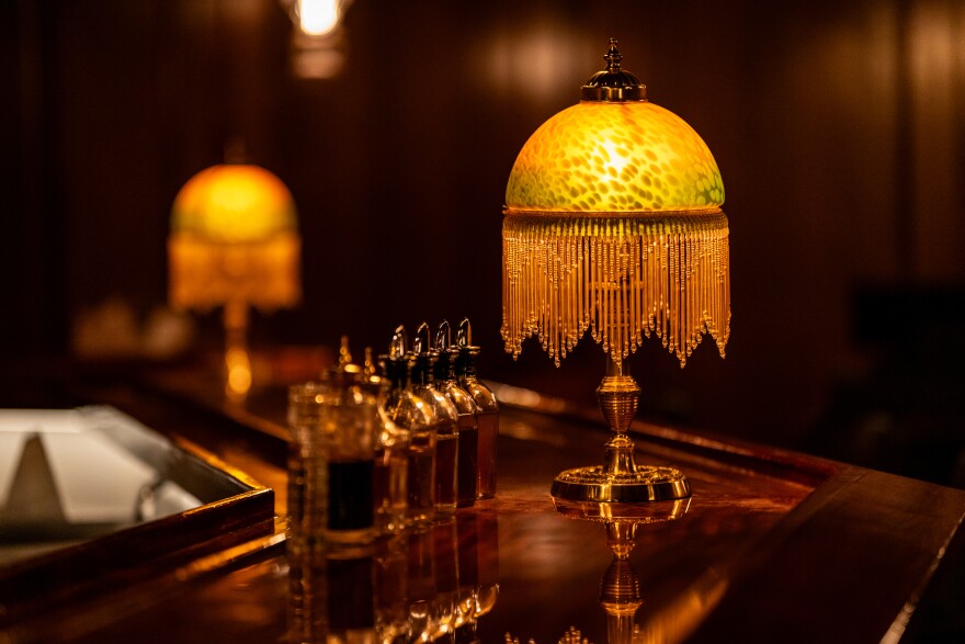 A dark, sophisticated wooden bar counter contains glass bottles filled with dark liquid and a glowing yellow lamp in an Art Deco style