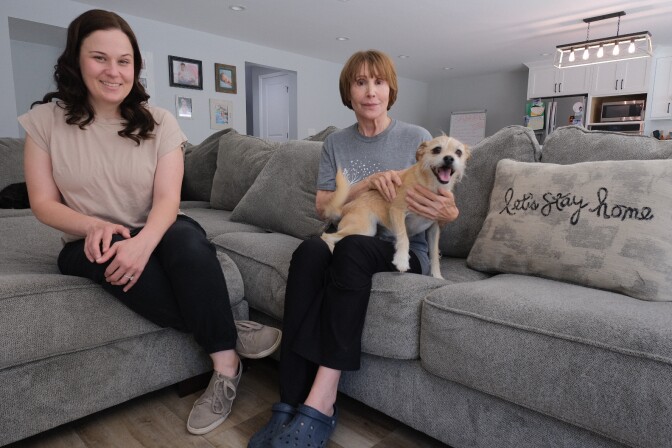 Two women sit on a grey couch in a living room and look at the camera. On the left smiling is a younger woman with dark hair down past her shoulders, wearing a tan shirt and black pants. In the center of the image is an older woman with light skin tone holding a happy dog. She has short auburn hair, a grey shirt and black pants.
