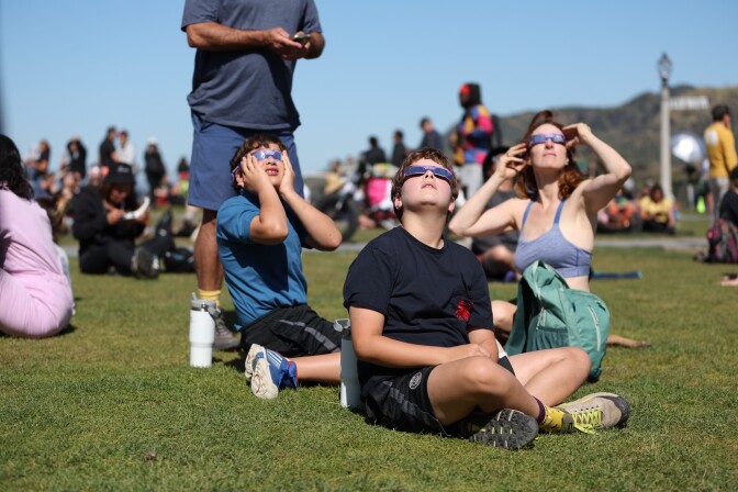 People sit on a lawn with special eyewear and look toward the sky.