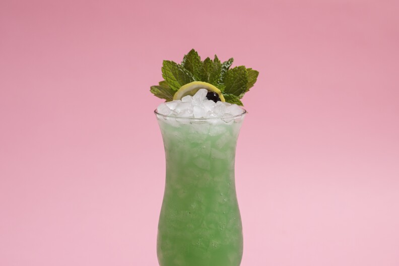a green cocktail in a glass with crushed ice and topped with mint leaves, set against a pink background