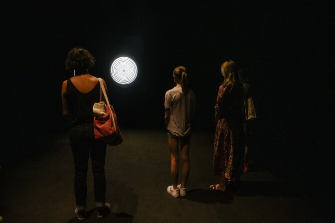 A spinning white and black disc, filled with spiral designs, hangs on the wall. A group of people stare at it in a black, darkened room.