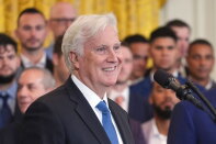 A man with white hair and a light skin tone smiles in a dark suit and blue tie, with a microphone in the foreground and a group of people in suits behind them.