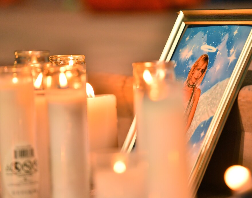A framed photo of Halyna Hutchins, with blond hair, a white dress, and a scarf, depicted with angel wings and a halo. The frame is surrounded by memorial candles burning.