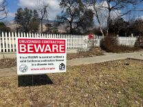 A red and white sign on a sidewalk reads "Unlicensed contractors BEWARE. It is a FELONY to contract without a valid California contractors license in a disaster area." 