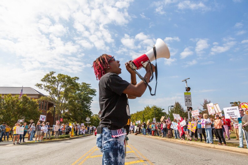 A Black man with a megaphone addresses people lining a street.