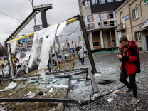 Rubble and shattered glass on the ground, a broken glass pane on the left, a street further to the left. Some people walk in the background; a young man in a red jacket is in the front right, looking at his phone.