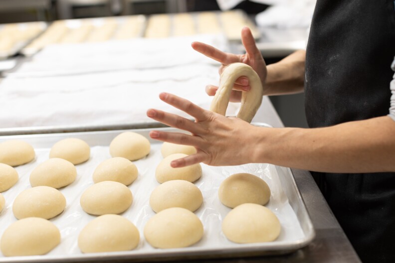A silver baking tray sits on a counter. On it is some white parchment paper, and perfectly evenly laid out rows of unbaked dough, slightly flattened spheres. A pair of light-skinned hands is pulling one of them into a bagel shape.