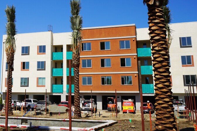 Yellow caution tape is tied in front of a building which is painted brown in the middle and beige on either side. The building has teal windows. Construction crews work on the building. 