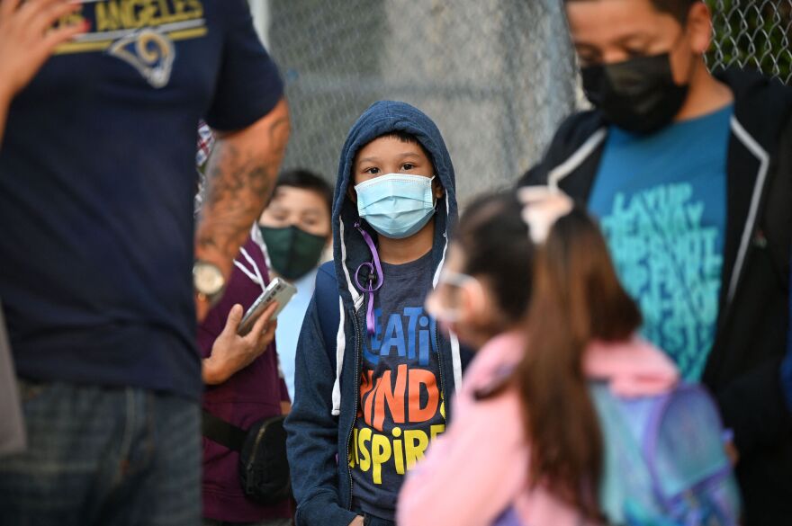 A student wearing a t-shirt, hooded sweatshirt and face mask stands among other students and parents near a chain-link fence.