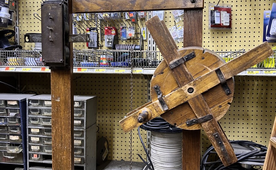 The interior of a packed hardware store aisle. Dozens of small products are hung up on the wall with metal pegs and long pieces of wire and cords can be seen on a shelf at the bottom. A wooden machine, with an aged metal fixture on the top left, is placed on the ground in front of the shelves.