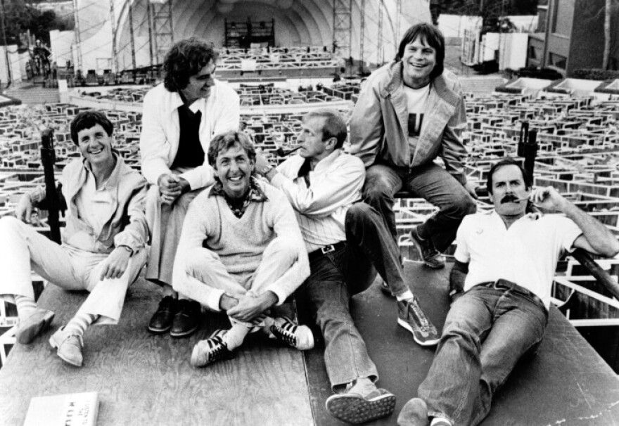 Six members of Monty Python in a black and white photo sit on plywood platform with the shell of Hollywood Bowl behind them.