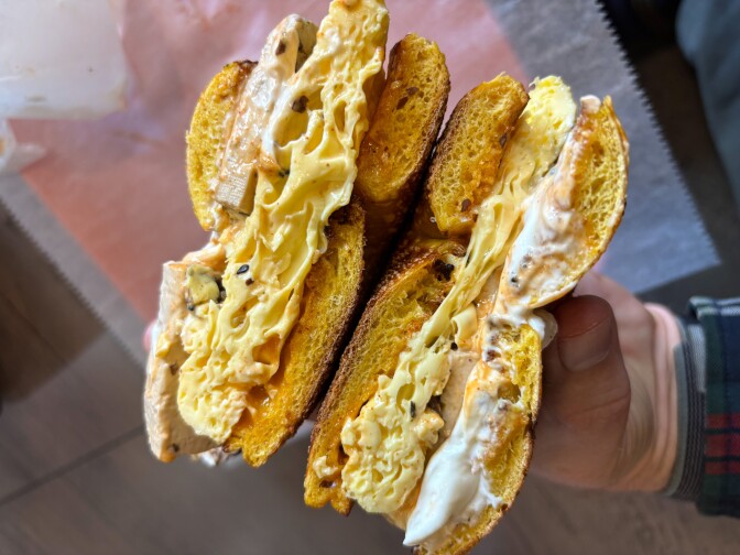 Handheld cross-section of a breakfast bagel sandwich showing folded soft scrambled eggs, melted cheese, marinated tofu, cream cheese spread, and a bright yellow egg bagel.