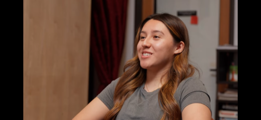 Side profile of young Latina with long brown hair with highlights, smiling, mid speech during an interview