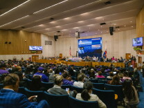 A large auditorium where a large crowd sits in blue chairs facing a dais with a large screen above. 