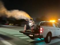A wide view look at a white moving truck as it drives into an intersection at night with a machine on the truck bed spraying insecticide into the air.