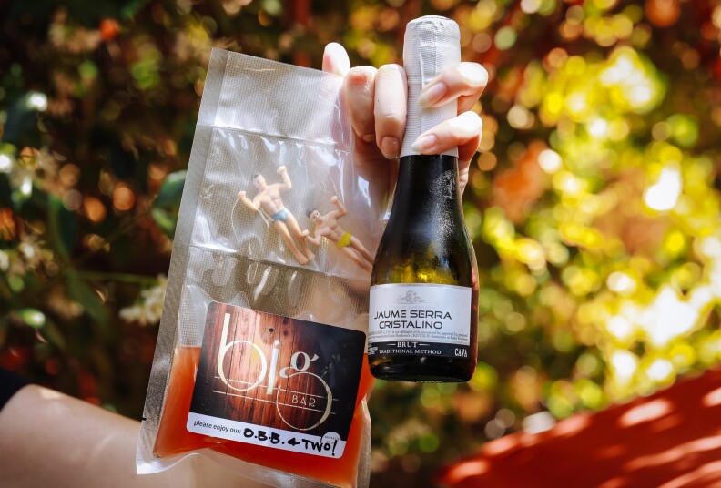 a hand holds a bag of liquid, a split of champagne and two plastic figurines