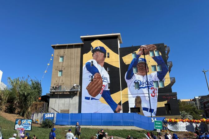 A mural featuring a baseball player on two different panels of a mural. A crowd is below looking at it.
