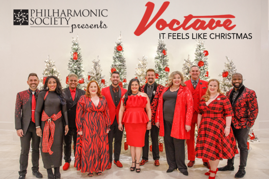 A promotional photo. The top left has the logo "Philharmonic Society presents" and the right of the photo reads "Voctave. It Feels Like Christmas." The photo features the Voctave a capella group standing in front of a beige-white background. The group features 11 men and women standing in front of 9 Christmas tress of varying heights. The Christmas trees are adorned with silvery tinsel and red decorations. The men and women are are varying heights and skin tones. There are 6 men and 5 women in alternating positions. They are all wearing festive suits and dresses with a mix of black and red colors. 