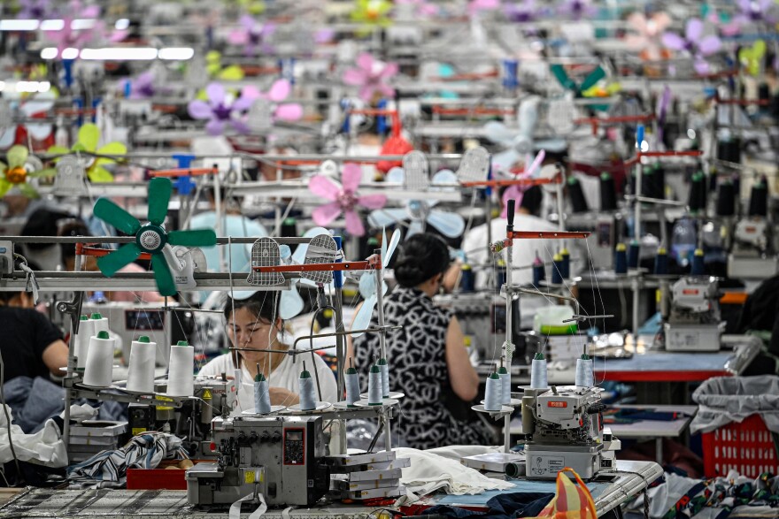 Women working in a textile factory. One woman with fair skin and dark brown or black hair wearing a white shirt is sitting down working on a garment. She's surrounded by machinery. The back of a woman with dark brown or black hair wearing a black and white colored giraffe print outfit is sitting down also working and surrounded by machinery.
