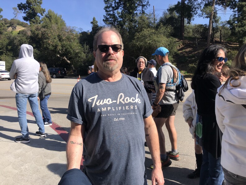 A light-skinned man with a thin goatee and thinning hair is wearing sunglasses and a gray t-shirt that reads Two-Rock Amplifiers. About eight other people are in the background in a parking lot.