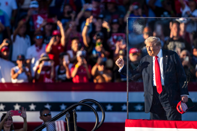 A man in a red tie and suit is gesturing to a crowd while on stage behind bulletproof glass at a political rally.