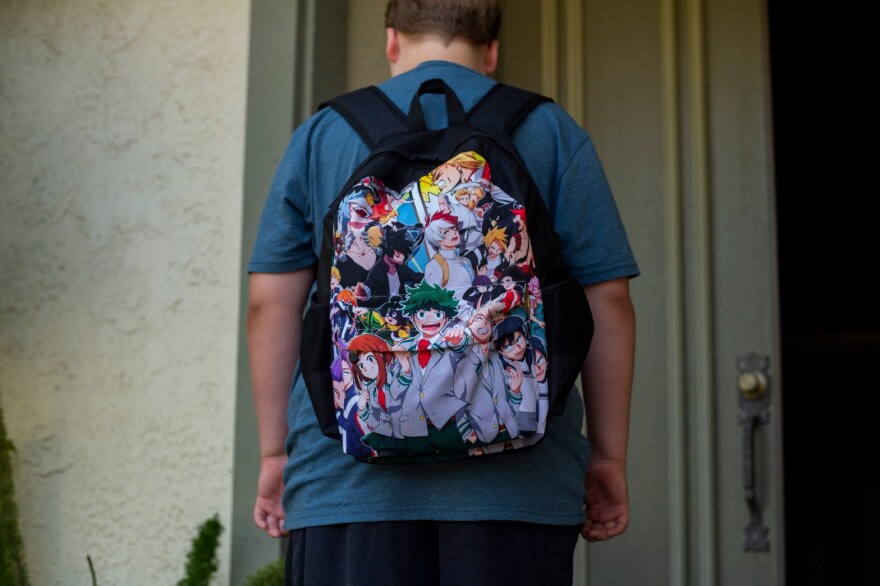 A student with his back to the camera wears a dark blue t-shirt and a backpack with anime characters.