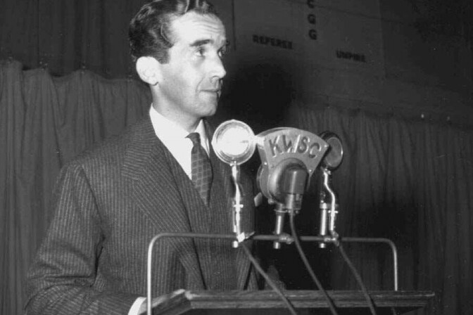 Black and white photo of a man wearing a suit speaking into a bank of microphones.