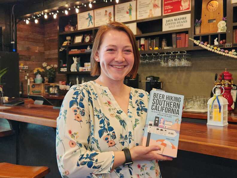 Author Johanna Flashman hold a copy of her book. The cover is bright blue with graphics of a beach, joshua tree and beer glasses. Flashman smiles wearing a white shift with blue flowers and stands in front of the wooden bar at RT Rogers