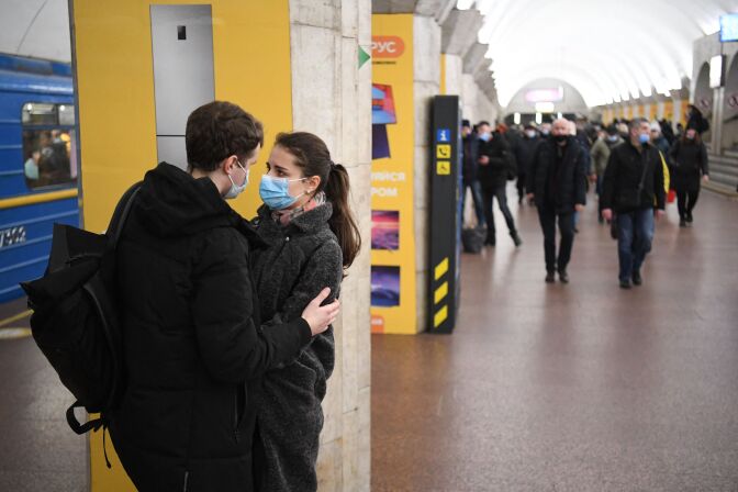 A man puts his arms on a woman in the lower left of the photo, holding her close. They stand in front of a yellow, metal, and stone column. They each wear dark jackets; her eyes look intense/concerned. A subway train is seen on the left, people walking down the platform to the right.