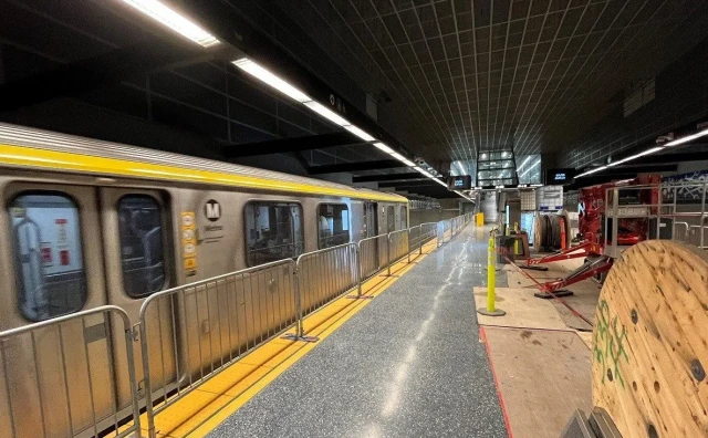 A semi-blurry silver Metro train runs along the lefthand side of the image. In the middle, there's a train platform whose edge is colored yellow. There are gates in between the platform and the train. On the righthand side are construction materials.
