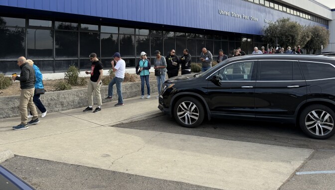 A line of people wait outside the Pasadena post office on Lincoln Ave. The building behind them is blue. 