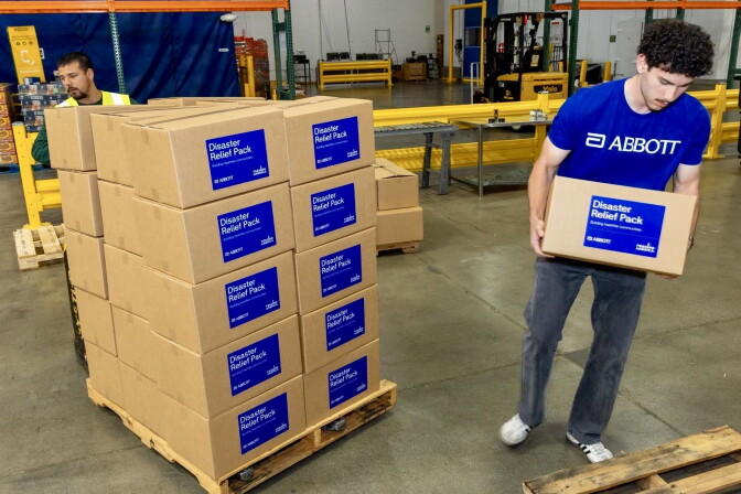 A male presenting person carries a cardboard box. A stack of about 30 boxes is next to him.