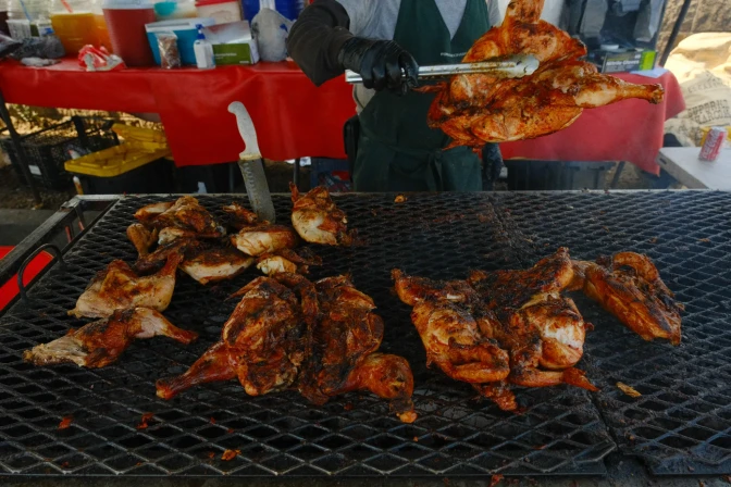 A vendor grills and flips seasoned chicken over an open flame at an outdoor food stand, with various supplies and drinks in the background.