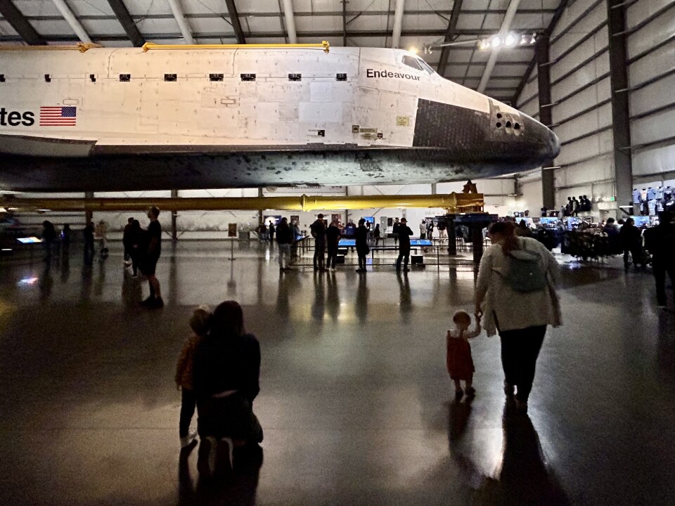 A mother kneels down next to her young child as they look towards a massive space shuttle with the words "Endeavour" facing the camera. Another mom is walking a few feet in front of them, holding an even younger child's hand.