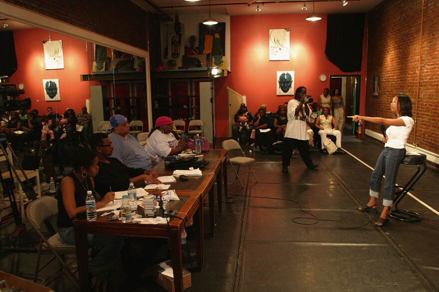 A young woman with medium brown skin, wearing a white top and denim jeans rolled up from the bottom, gestures during an audition before a panel of people seated at a long wooden table.