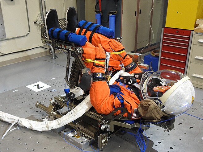 A mannequin is positioned on its back in an orange jumpsuit and helmet.