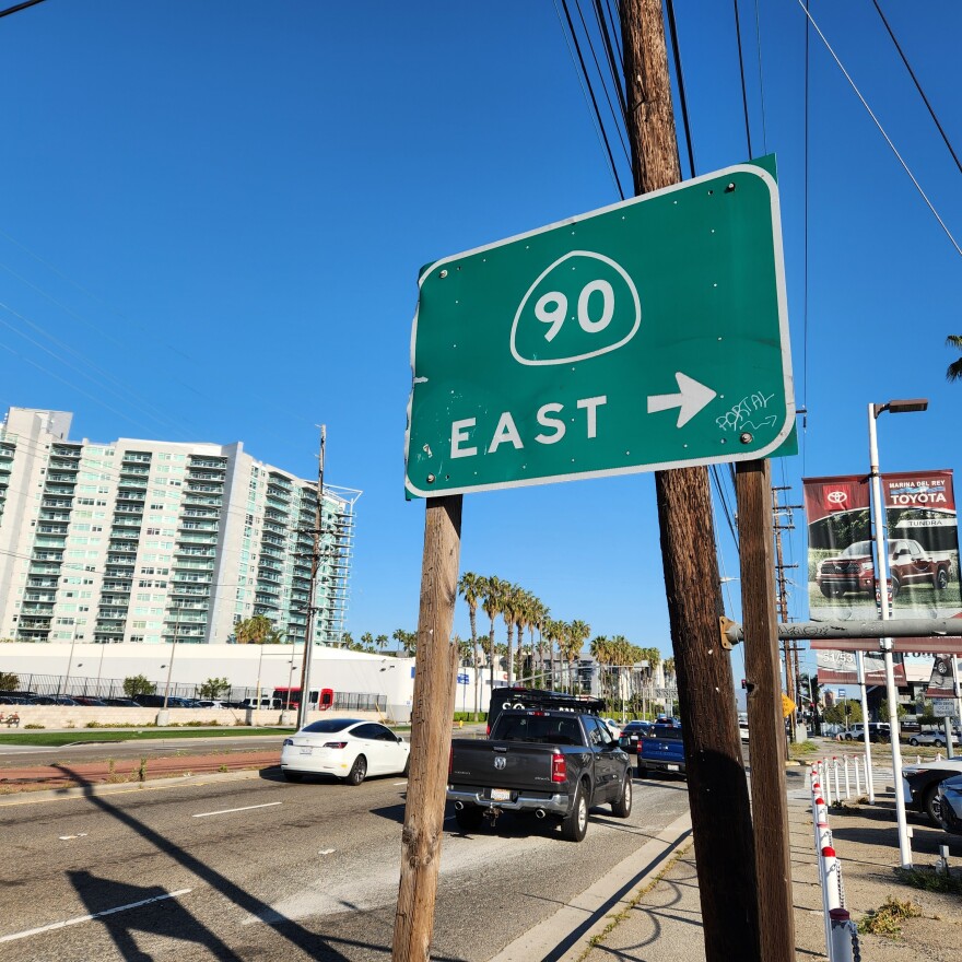A green street sign denoting the 90 Freeway entrance, with the word "East" and an arrow below it in white. In the background is a highway with palm trees and a blue sky.