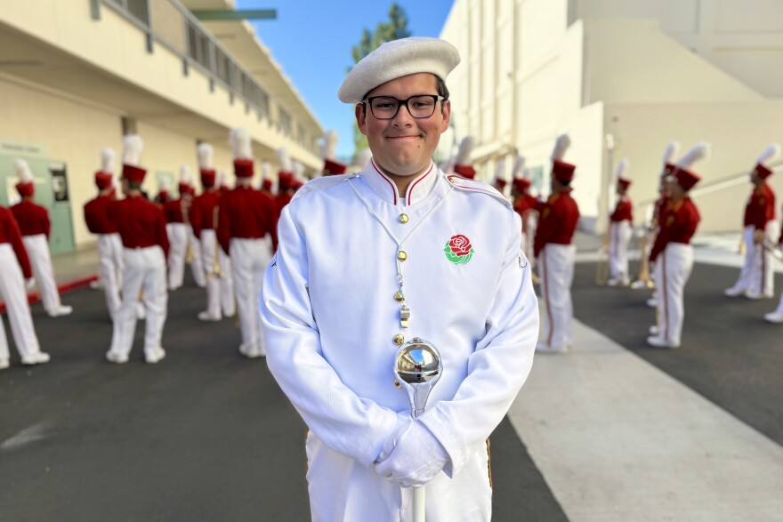 A man with medium-light skin tone smiles for a portrait. He wears a pristine white uniform with a red rose on the lapel and in his hands he holds a mace.