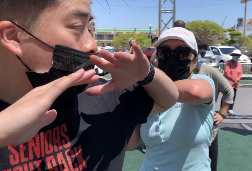 An older Vietnamese American woman wearing a mask, sunglasses and a light blue blouse practices self-defense moves on a male volunteer wearing a mask and a black t-shirt outside the Asian Garden Mall in Westminster.